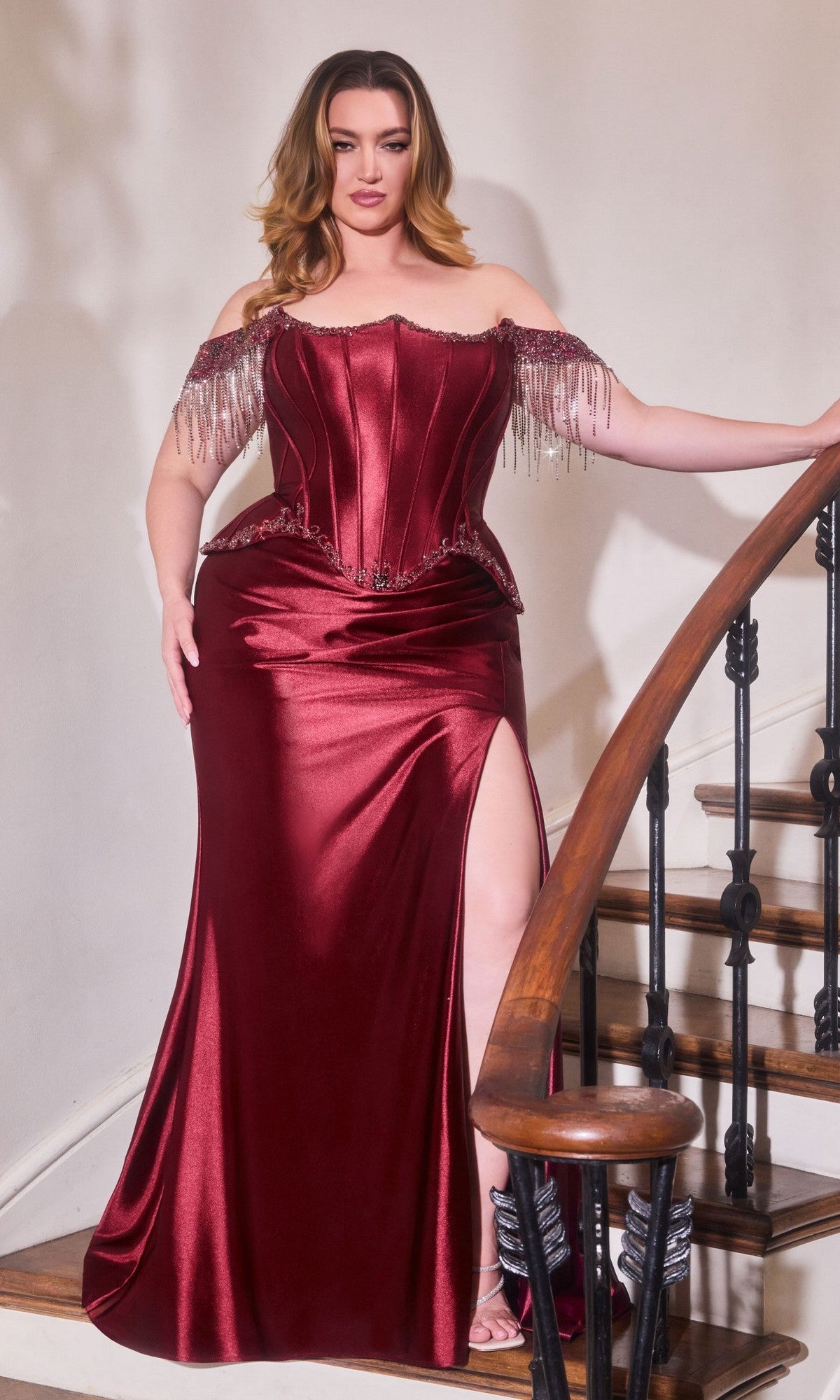 A woman wearing the Ladivine CD688 long prom dress in burgundy, featuring an off-shoulder satin corset design with a thigh-high slit, stands on a wooden staircase holding the railing.